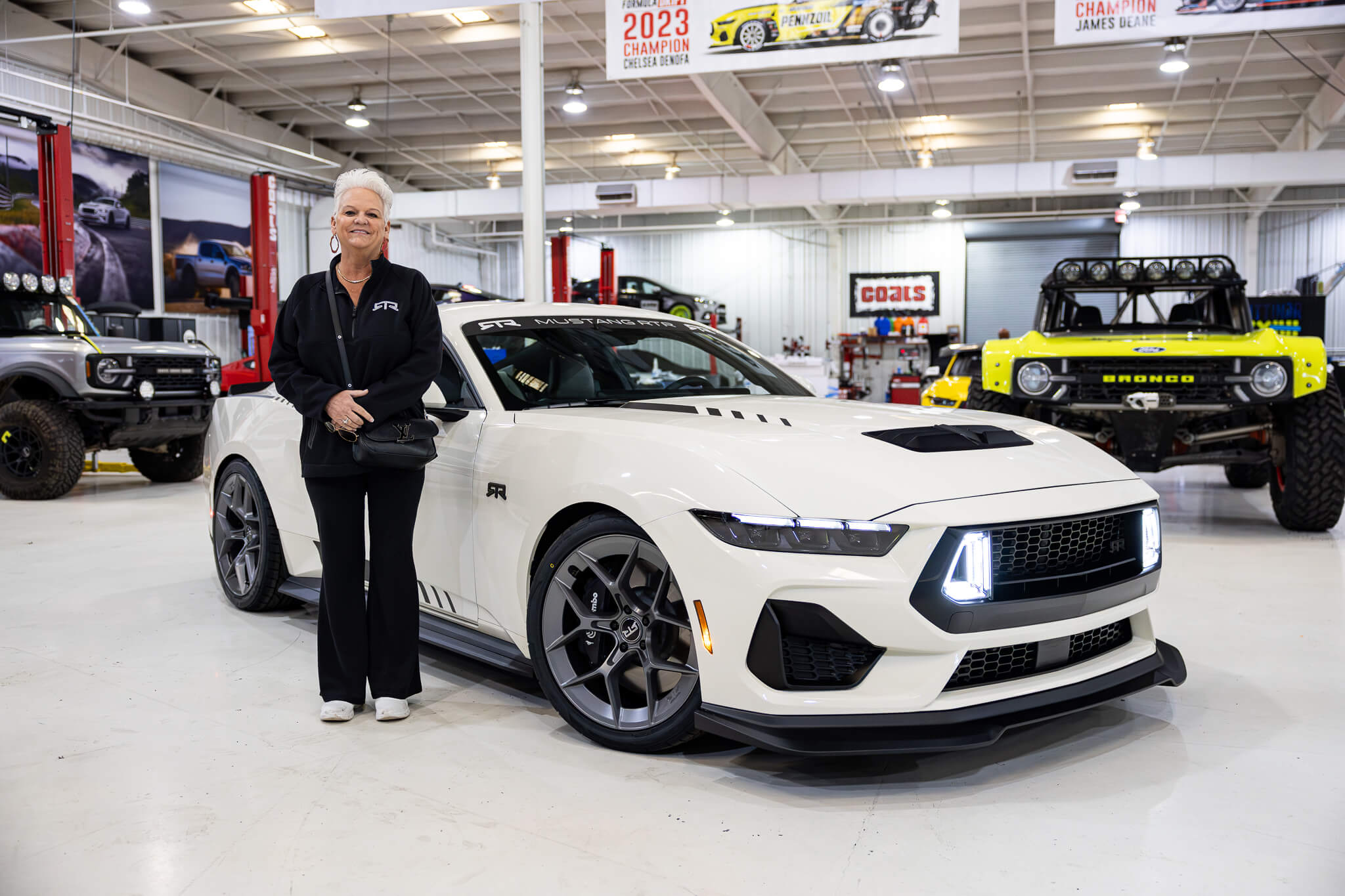 A woman in an RTR jacket stands with her 2025 Wimbledon white 60th anniversary Mustang RTR inside the RTR shop