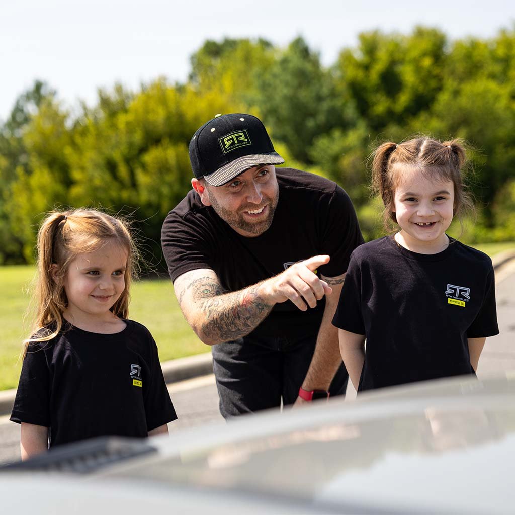 Man in black RTR hat pointing at two girls smiling, wearing the RTR Spec 5 10th Anniversary Tee, celebrating RTR's Ready to Rock spirit.